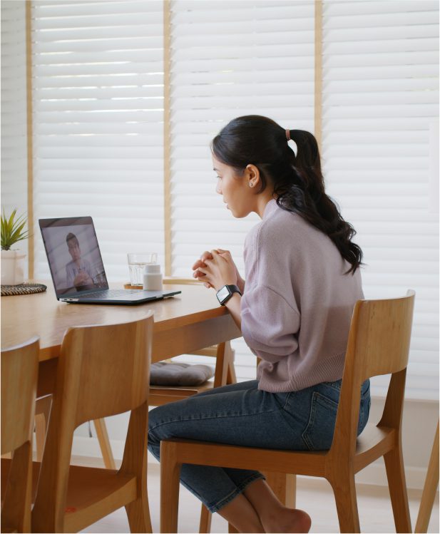 A woman sits at a table using a laptop for a video call. She has a ponytail and is wearing a lavender sweater and jeans. Blinds cover the window behind her.