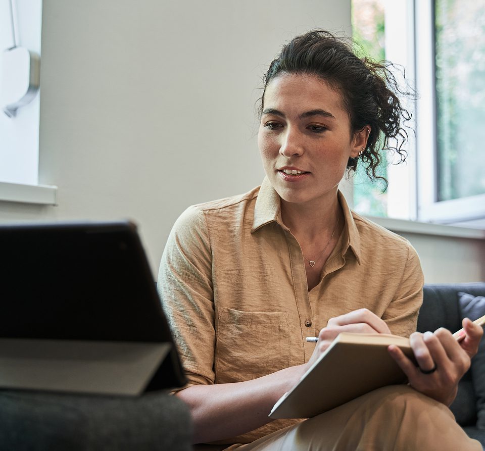 Woman in a beige shirt sits on a sofa, holding a notebook and pen, looking at a tablet on a stand.
