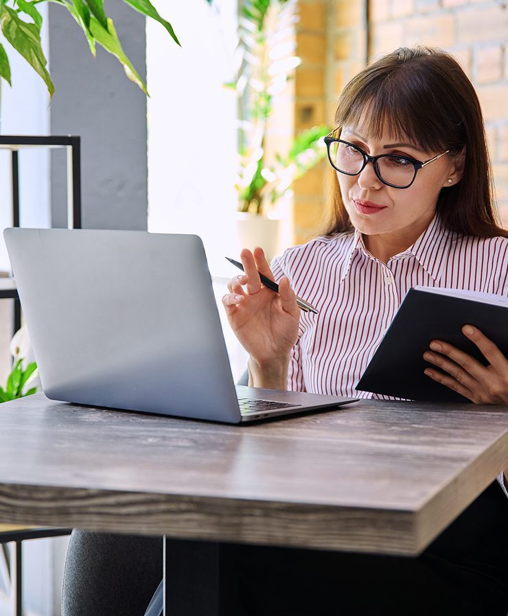 Woman with glasses holding a tablet and pen, sitting at a table, looking at a laptop screen.